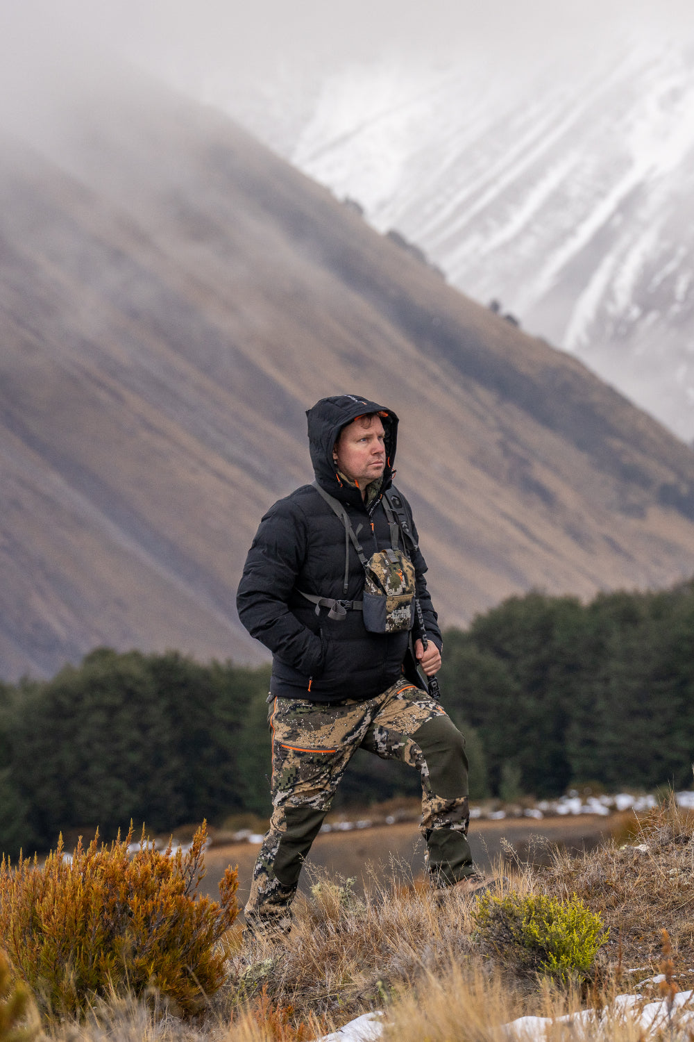 Person in camouflage clothing and black jacket standing in a mountainous landscape with snow-capped peaks.