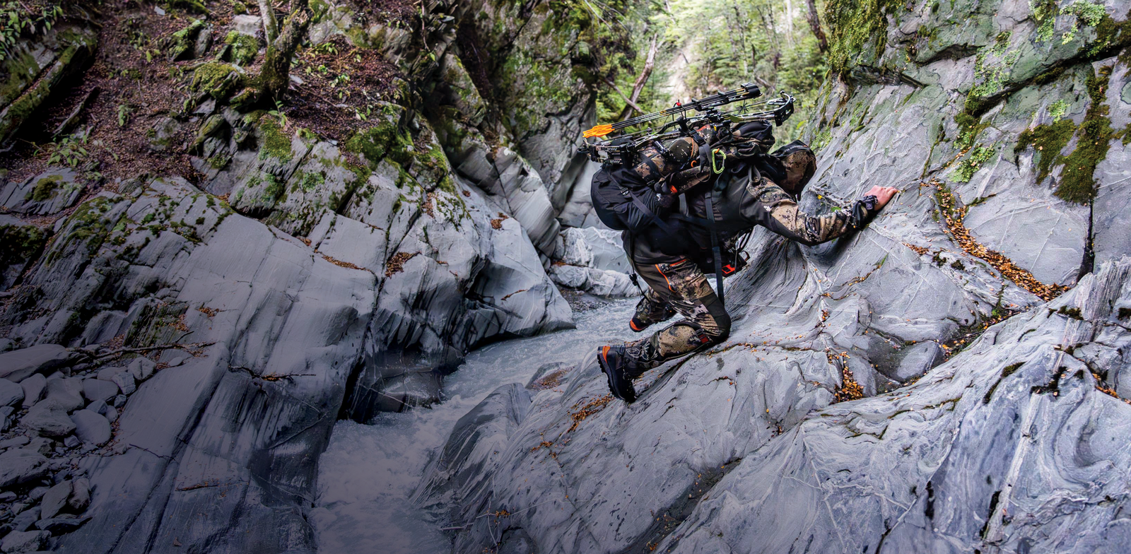 Person in camouflage gear climbing rocky terrain with backpack and equipment.