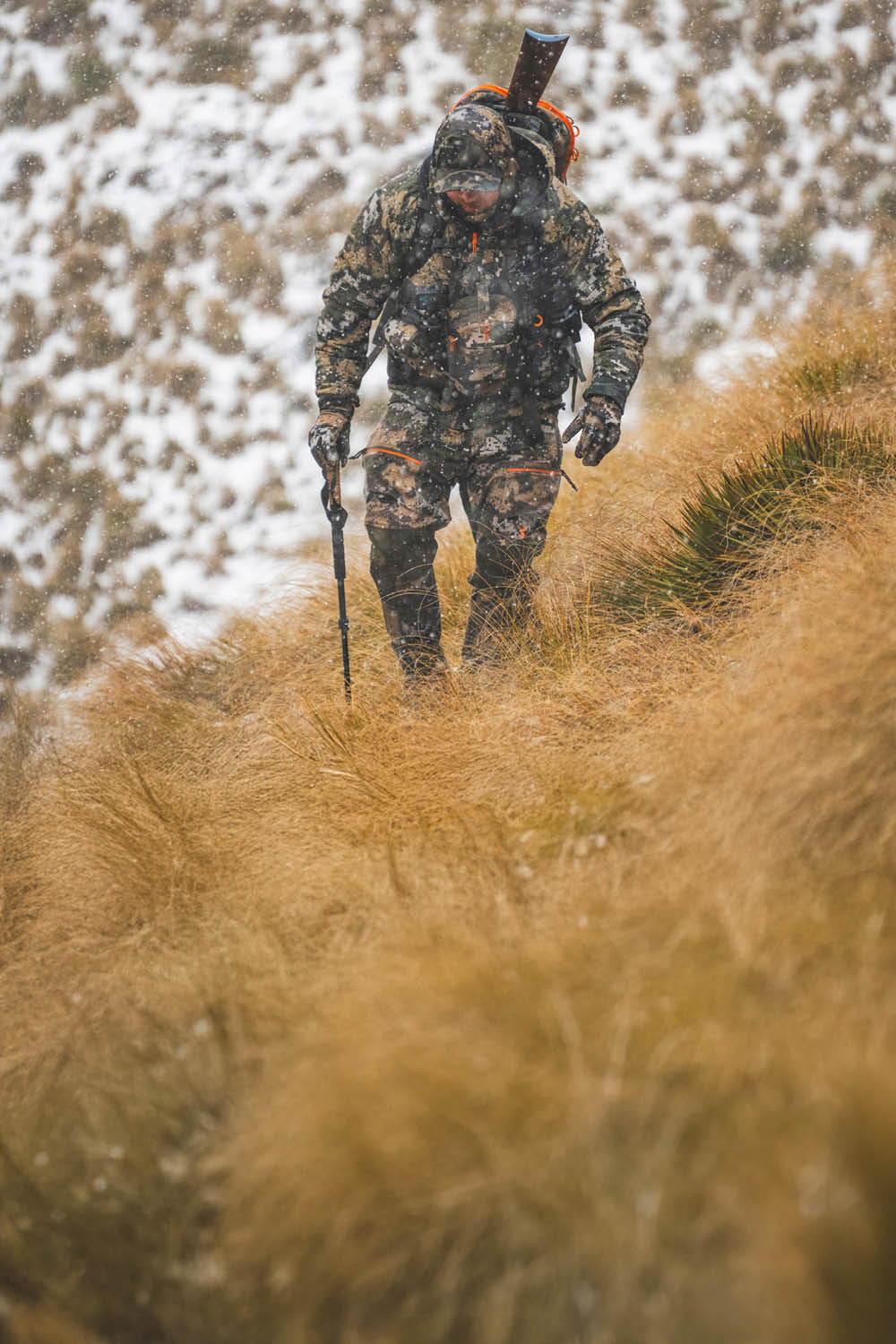 Hunter in camouflage walking through tall grass with a bow, surrounded by snow.