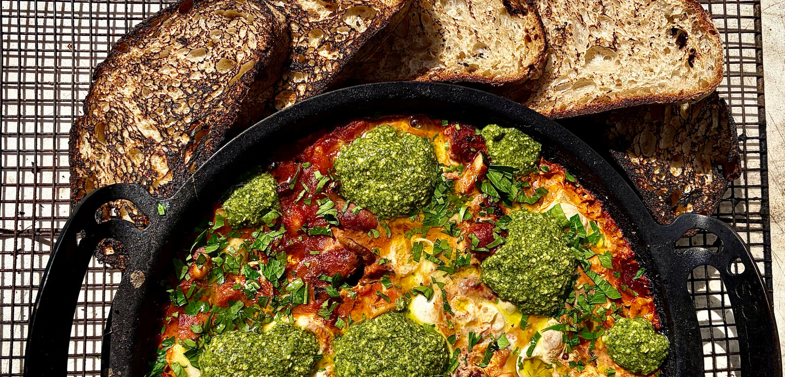 Dish with green sauce and vegetables in a cast iron skillet surrounded by bread on a cooling rack.