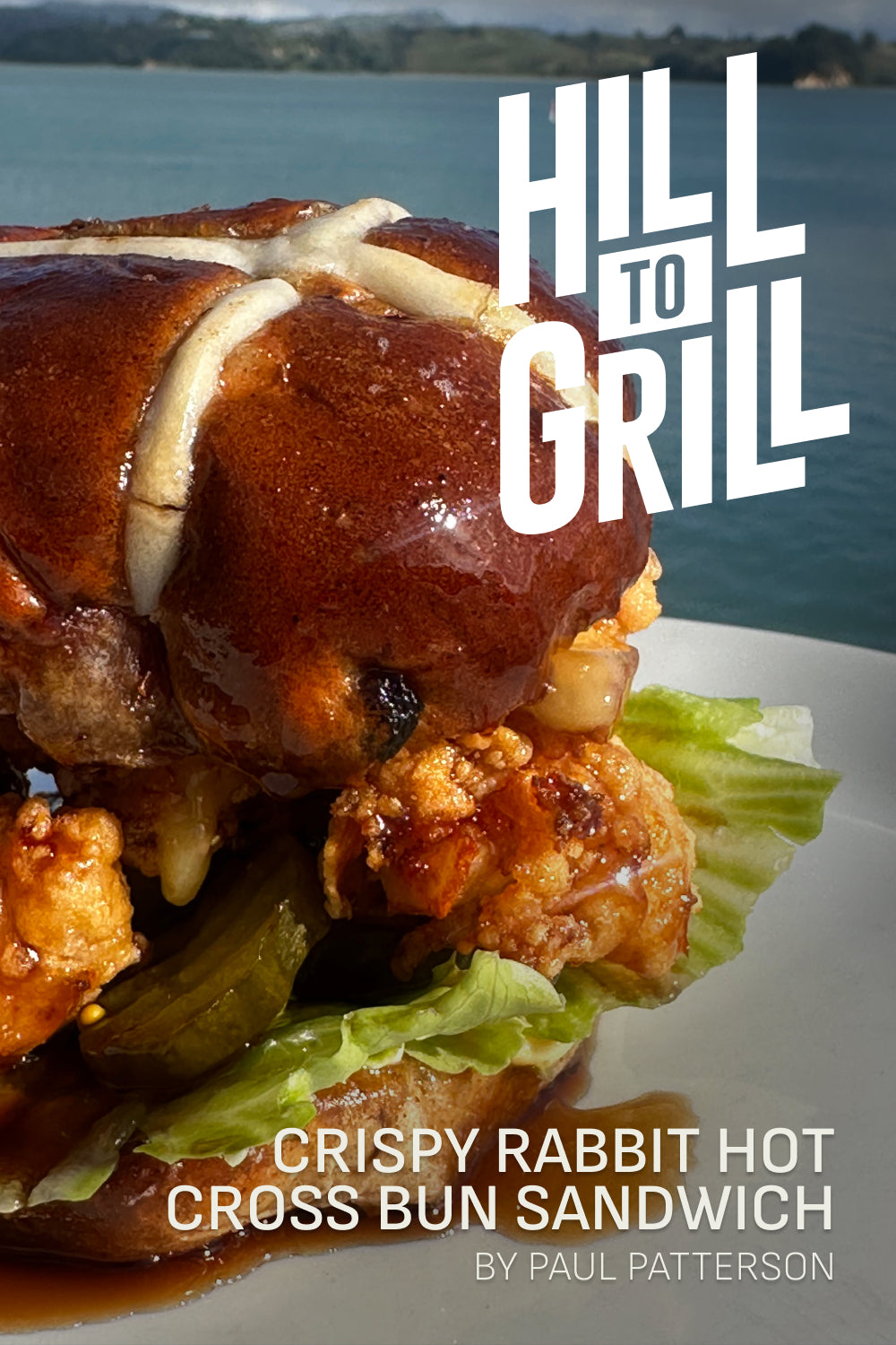 Crispy rabbit hot cross bun sandwich on a plate with a scenic background, featuring the text 'Hill to Grill' and 'Crispy Rabbit Hot Cross Bun Sandwich by Paul Patterson'.